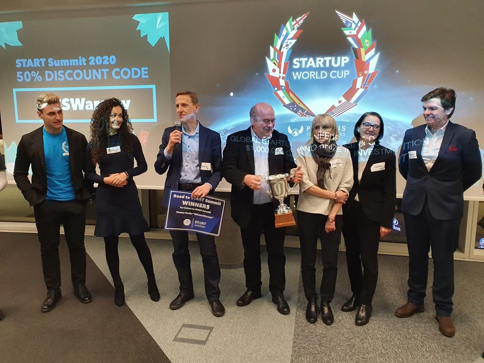 na zdjęciu widać siedem osób, trzy kobiet i czterech mężczyzn, w różnym wieku, stojących w sztucznie oświetlonym pomieszczeniu. Za nimi widać ekran z angielskojęzycznym napisem  Startup World Cup. Jedna z osób trzyma w ręku puchar