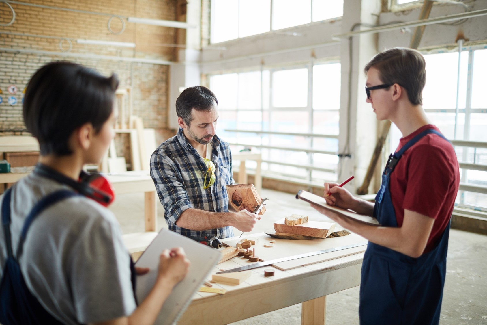 Zdjęcie przedstawia pracownie stolarską. Stolarz pokazuje młodym uczniom, jak odpowiednio przygotować drewno 